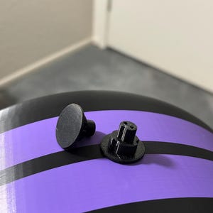 May include: Two small, black plastic components. One is a flat disc with a stem, the other has a cylindrical base with a textured top. The items are on a surface with black and purple stripes.