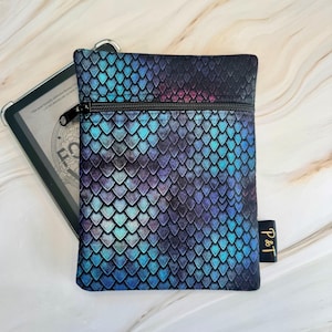 May include: A small, rectangular pouch with a zipper closure, featuring a scale-like pattern in shades of blue, purple, and teal. The pouch has a black zipper and a small black tag with gold text. An e-reader is partially visible in the background.