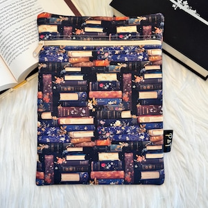 May include: A rectangular fabric pouch featuring a dark navy blue background with a repeating pattern of stacked books in shades of blue, red, and cream, accented with small floral details. The pouch has a cream-colored zipper and a black side tag.