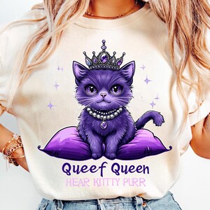 May include: Cream-colored t-shirt featuring a purple cartoon cat wearing a crown and pearl necklace, sitting on a purple pillow. The text "Queef Queen" and "Hear Kitty Purr" are printed below the cat.