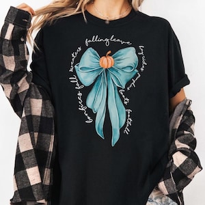 May include: Black t-shirt featuring a teal bow with an orange pumpkin in the center. The shirt has the words "falling leaves, cozy fires, fall sweaters, pumpkin boots, football, hayrides" in a circular design around the bow.