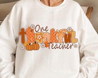 May include: White sweatshirt with the text "One Thankful Teacher" in orange and brown lettering. The design includes pumpkins, daisies, and fall leaves, perfect for the autumn season. A cozy and festive top for educators.