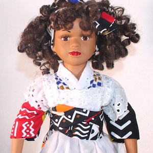 Puede incluir: Muñeca con piel morena, cabello castaño rizado y lápiz labial rojo. La muñeca viste una blusa calada blanca con una faja estampada y una falda blanca. Un lazo estampado azul, negro y rojo adorna el cabello de la muñeca.