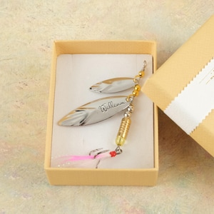 May include: A silver fishing lure set, engraved with the name "William", presented in a beige gift box. The lures have a reflective surface and are adorned with gold and silver beads, and pink and white feathers.