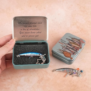 以下が含まれることがあります： ルアーが入った、水色の金属製の箱と「母はいつも人生はチョコレートの箱のようなものだと言っていました。何が出てくるかわからない」という引用。魚のイラストが描かれた2つ目の箱も表示されています。