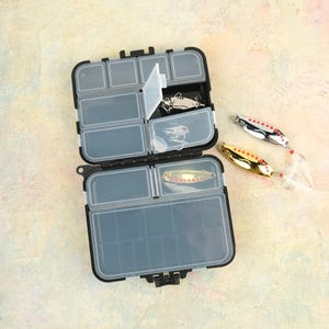 K&ouml;nnte beinhalten: Eine offene, schwarze und transparente Angelkiste mit mehreren F&auml;chern. Drei Angelk&ouml;der sind zu sehen: einer goldfarben und zwei silberfarbene, jeweils mit roten Akzenten und wei&szlig;en Federenden. Die Box befindet sich auf einer hellen Oberfl&auml;che.