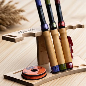 以下が含まれることがあります： 3本の釣り竿とオレンジ色の釣り糸のスプールが入った木製の釣り竿ホルダー。 竿は木製のハンドルと色のアクセントが付いています。 ホルダーには「Alexander」という名前が刻まれています。