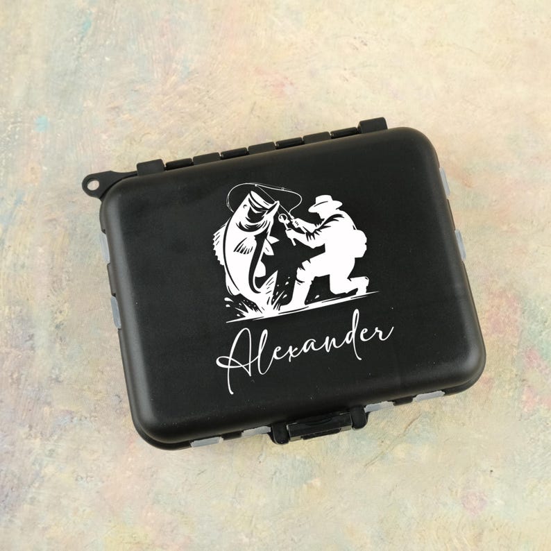 K&ouml;nnte beinhalten: Schwarze Angelkiste mit einer wei&szlig;en Grafik eines Fischers, der einen Fisch f&auml;ngt. Der Name "Alexander" ist darunter in Kursivschrift geschrieben. Die Box hat einen Klappdeckel und einen sicheren Verschluss.