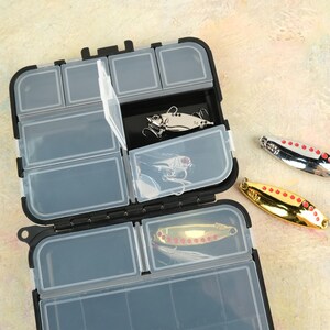 K&ouml;nnte beinhalten: Eine schwarze Kunststoff-Angelbox mit transparenten F&auml;chern, ge&ouml;ffnet, um Angelk&ouml;der zu zeigen. Die K&ouml;der sind silber- und goldfarben, mit roten Akzenten. Die Angelbox befindet sich auf einer hellen Oberfl&auml;che.