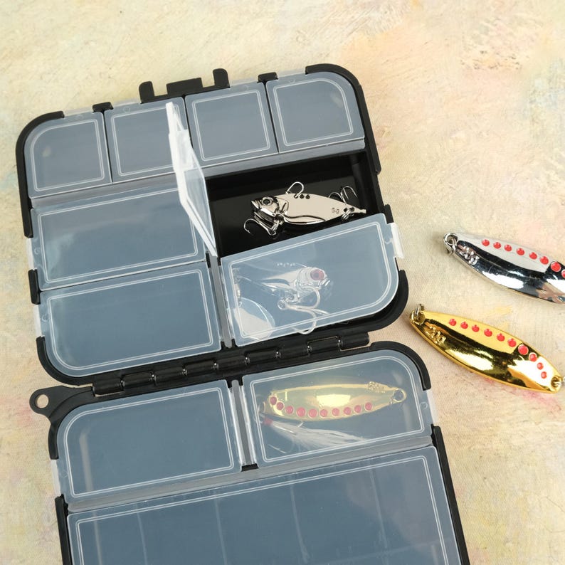 K&ouml;nnte beinhalten: Eine schwarze Kunststoff-Angelbox mit transparenten F&auml;chern, ge&ouml;ffnet, um Angelk&ouml;der zu zeigen. Die K&ouml;der sind silber- und goldfarben, mit roten Akzenten. Die Angelbox befindet sich auf einer hellen Oberfl&auml;che.