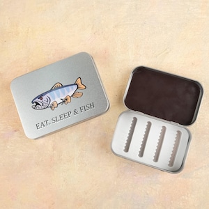 以下が含まれることがあります： 漫画の魚のイラストと「EAT. SLEEP & FISH」の文字が入った、銀色の金属製のフライフィッシングボックス。ボックスは開いており、フライを保持するためのフォームインサートが付いた白い内部が見えます。ふたは左側に閉じています。