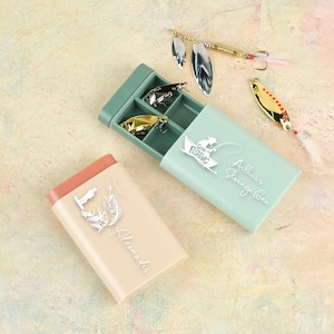 May include: A light green fishing tackle box with three lures inside, and a peach-coloured box with the name "Alexander" on it. The open box has the words "Go Fishing" and "Arthur's Storage Box" on it. Several fishing lures are scattered around.