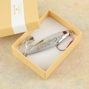 以下が含まれることがあります： 「I LOVE YOU MORE THAN YOU LOVE FISHING」の文字とハートマークが入った銀色のルアーが、ベージュ色のギフトボックスに入っています。ルアーには赤い宝石とフックが付いています。ギフトボックスには「JUST FOR YOU」と書かれた白いリボンが付いています。