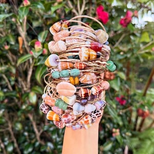 May include: A collection of gold bangles, each decorated with seashells and colourful beads. The bracelets display a variety of natural shell shapes and beads in green, orange, and pink. The background is a vibrant green with touches of red flowers.