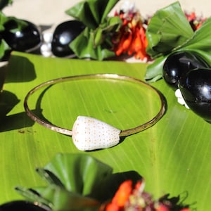 May include: A gold-colored bangle bracelet with a white seashell accent, resting on a large green leaf. The bracelet is surrounded by black, glossy objects wrapped in green leaves and red flowers.