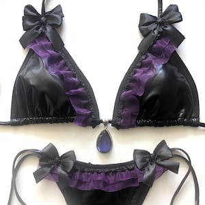 May include: Black lingerie set with a triangle top and thong bottom. The top features purple ruffled trim, black bows, and a teardrop-shaped pendant. The thong also has purple ruffles and black bows.