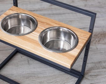 May include: A modern pet feeder with two stainless steel bowls set into a light wood platform. The platform is supported by a dark gray metal frame. The bowls are designed for food and water.