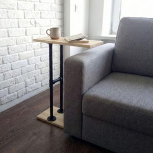 Puede incluir: Pequeña mesa auxiliar de madera clara con patas de tubo metálico negro. Una taza beige y un libro descansan sobre la mesa. La mesa está colocada junto a un sofá gris. Sus dimensiones son de unos 30 cm de ancho y 61 cm de alto.