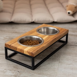 May include: A wooden pet bowl stand with two stainless steel bowls. The stand is made of light brown wood and has a black metal frame. One bowl is filled with pet food.