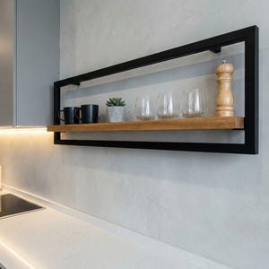 May include: A rectangular floating shelf made of black metal and wood, displaying kitchenware. It features two black mugs, a small plant, four glasses, and a wooden pepper mill. The shelf is mounted on a light grey wall.