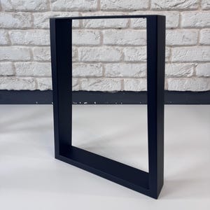 May include: A black, rectangular table leg frame. The frame is made of solid material and has a matte finish. The table leg is angled slightly, showcasing its geometric design. The background features a white brick wall.