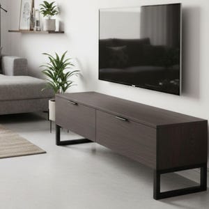 Puede incluir: Un mueble de TV de madera marrón oscuro con dos cajones y patas de metal negro. El mueble está situado debajo de un televisor de pantalla plana grande montado en una pared blanca. Un sofá gris y plantas son visibles en el fondo.