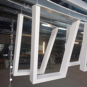 May include: White painted metal table legs hanging in a workshop. The legs have a modern, geometric design, with a rectangular top and angled supports. The workshop has a concrete floor and overhead lighting. The legs are likely for a table.