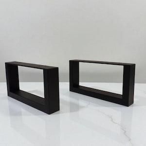 May include: Two rectangular, matte black table legs. Each leg is a hollow, box-like shape with a clean, modern design. The legs are positioned on a white surface, suggesting a minimalist aesthetic.