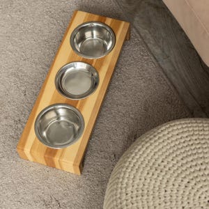 May include: A wooden pet feeder with three stainless steel bowls. The feeder is made of light-coloured wood with a striped pattern. The bowls are evenly spaced and appear to be empty. The feeder is designed to sit on the floor.