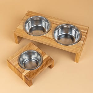 May include: Two wooden pet food bowls with stainless steel inserts. One bowl is a single, square design, and the other is a double bowl design. The wooden bases are light brown with visible wood grain. The background is a solid tan colour.
