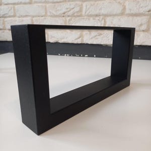 May include: A black rectangular shelf with a hollow centre. The shelf has a matte finish and a simple, modern design. It is displayed against a white brick wall. The shelf's dimensions are not visible.