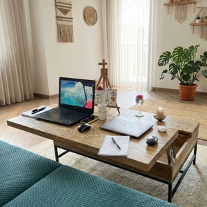 May include: A wooden coffee table with a lift-top design, featuring a laptop, mouse, and notepad. The table is set in a living room with a laptop, mouse, and notepad. The room has a plant, artwork, and a window.