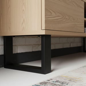 May include: Close-up of a light wood cabinet with a rectangular black metal base. The cabinet has a natural wood grain finish. The black metal legs are square and provide a modern, minimalist aesthetic. The cabinet is set against a white brick wall.