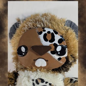 May include: A handmade plush toy with a brown and white face, featuring large black eyes and a stitched design. The toy has a furry, light brown body and a white and black patterned chest.