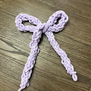 May include: A lavender-colored, braided bow made of a soft, plush material. The bow is tied in the center and has two long, dangling ends. The bow is lying on a wooden surface.