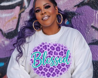 May include: A white long-sleeve sweatshirt featuring a purple leopard print circle with the word "Blessed" in teal script. The person in the image is wearing gold hoop earrings and a gold chain necklace.