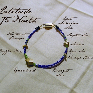 以下が含まれることがあります： 銀と木製のビーズが付いた青と緑のビーズブレスレット。ブレスレットには「Latitude 75' North」というテキストと、北極圏の水域と陸地の名前が書かれています。