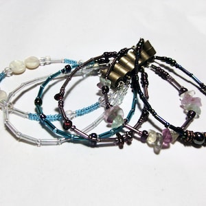 May include: A set of five beaded bracelets featuring a mix of white, blue, purple and green beads. The bracelets are layered on top of each other and feature a variety of bead shapes and sizes.