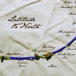 May include: A beaded necklace with blue and green beads, representing the 15th parallel north. The necklace includes labels for locations along the line, including Da Nang, Bangkok, Manila, Goa, Anantapur, and the Arabian Sea. The text "Latitude 15' North" is written at the top of the image.