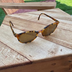 Może przedstawiać: Okulary przeciwsłoneczne w kolorze żółwia z brązowymi soczewkami leżą na drewnianym stole. Oprawki mają nakrapiany wzór w odcieniach brązu i bursztynu. Soczewki są w ciepłym brązowym kolorze, oferując estetykę vintage.