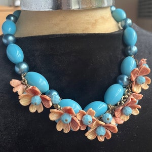 Puede incluir: Un collar de cuentas turquesa y melocotón con un diseño floral. El collar está hecho de cuentas redondas grandes de color turquesa y cuentas más pequeñas de color melocotón que tienen forma de flores. Las flores están agrupadas en el centro del collar.