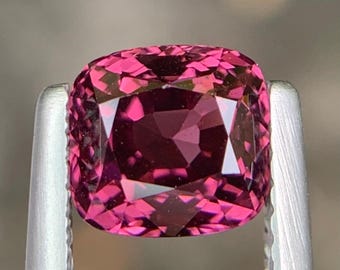 May include: A cushion-cut, faceted gemstone held by silver tweezers. The gem is a deep magenta color with multiple facets reflecting light. The gemstone's shape is square with rounded corners, showcasing its brilliance.