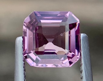 May include: A rectangular-cut gemstone in shades of lavender and purple, held by silver tweezers. The gem's facets reflect light, highlighting its clarity and colour. This gemstone is likely for jewellery making or collecting.