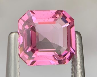 May include: A rectangular-cut pink gemstone held by silver tweezers. The gem has a vibrant, light pink hue with multiple facets that reflect light. The gemstone is a square shape with beveled edges.