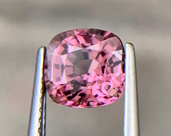 Op de afbeelding: Een gefacetteerde, kussenvormige roze edelsteen, vastgehouden door een zilveren pincet. De edelsteen heeft een zachte roze tint met interne facetten die licht reflecteren. De pincet is scherp, wat de helderheid en kleur van de edelsteen benadrukt.