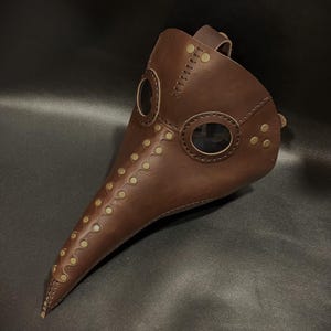 May include: A brown leather plague doctor mask with brass accents and two clear eye pieces. The mask has a long, pointed beak and is secured with a leather strap.