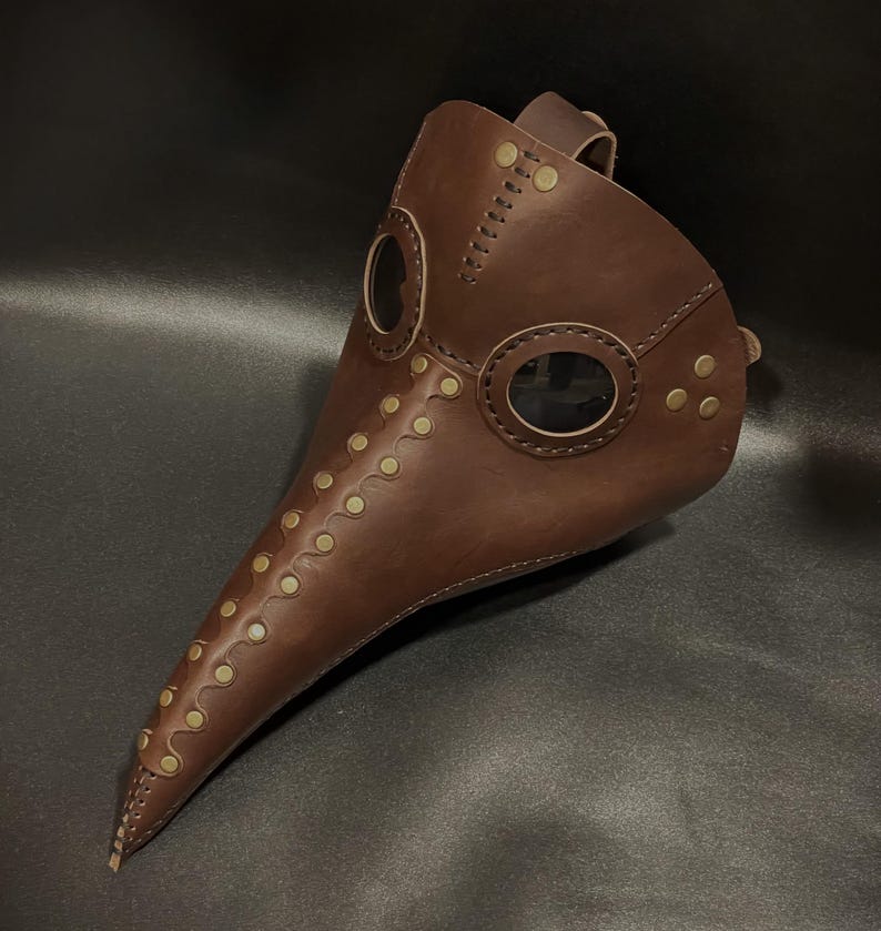 May include: A brown leather plague doctor mask with brass accents and two clear eye pieces. The mask has a long, pointed beak and is secured with a leather strap.