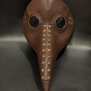 May include: A brown leather plague doctor mask with a long beak and two round eyeholes. The mask is decorated with brass rivets and stitching.