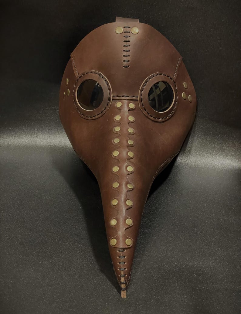 May include: A brown leather plague doctor mask with a long beak and two round eyeholes. The mask is decorated with brass rivets and stitching.