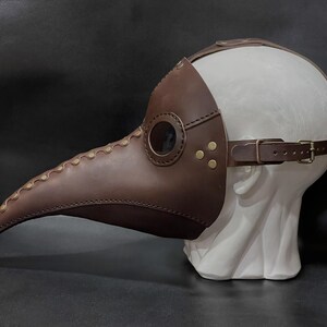 May include: A brown leather plague doctor mask with a long beak and eye holes. The mask is on a white mannequin head.